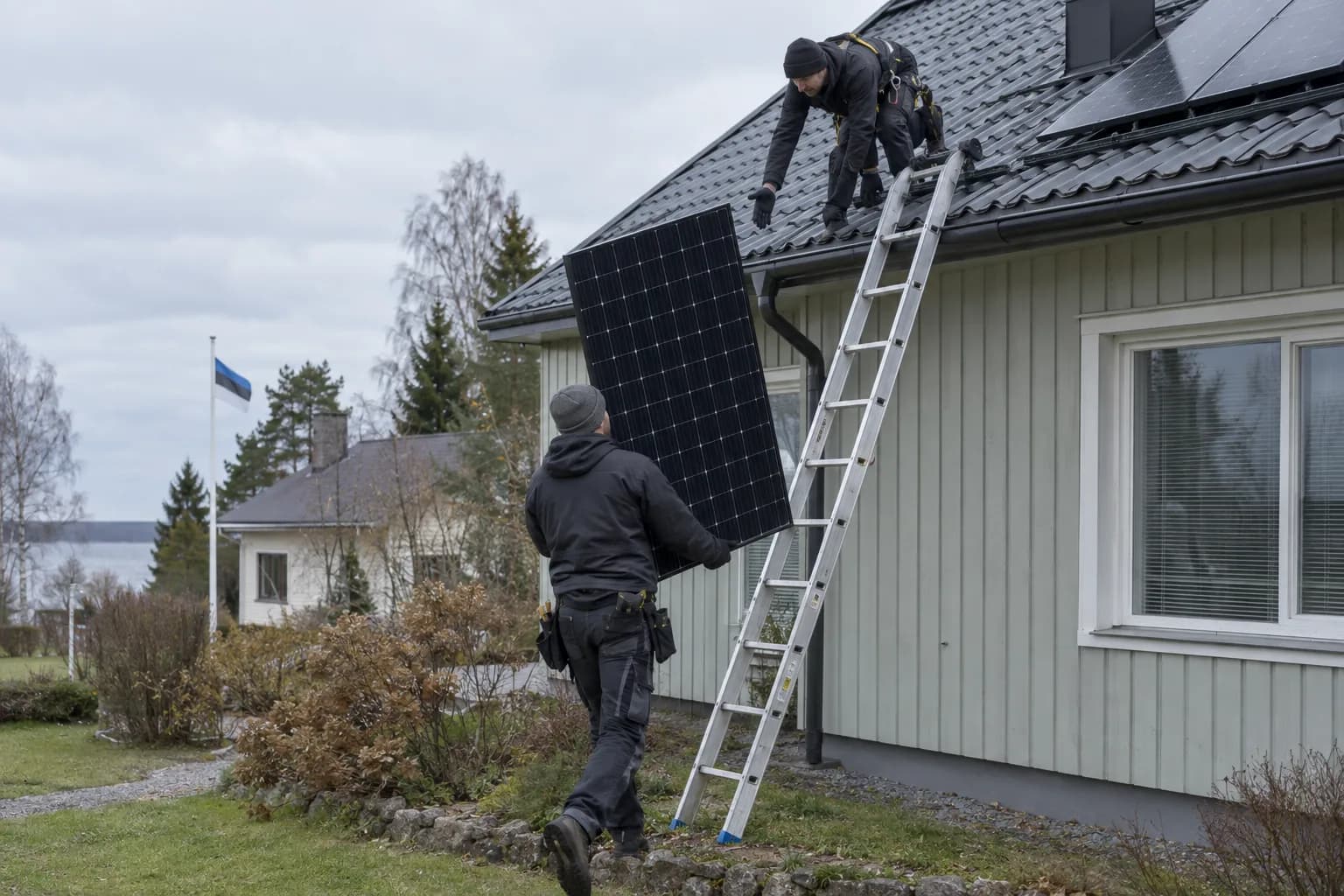 Paigaldaja kannab päikesepaneeli redeli juurde, teine töötaja ootab katusel